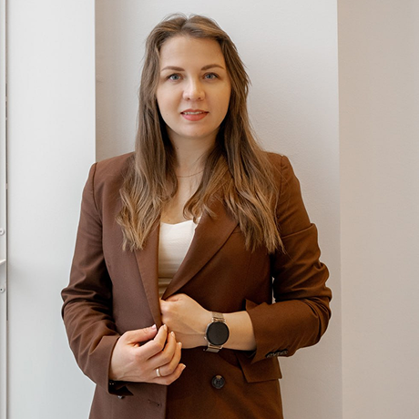 Professional woman stands in a doorway wearing a brown blazer and white top, looking at the camera with a neutral smile, hands lightly touching the blazer lapel.